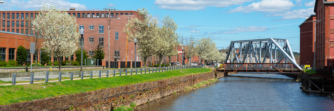 Pemberton Mill Bridge on the Merrimack River North Canal at Lawrence Heritage State Park in downtown Lawrence, Massachusetts MA, USA. The bridge is a steel truss bridge with a pedestrian walkway and a view of the river and city skyline. The image is taken during the day with clear skies and vibrant colors, showcasing the historic architecture and natural beauty of the area.