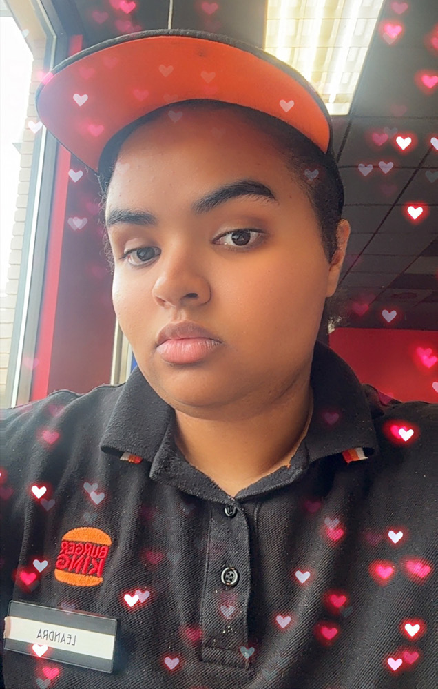 A Black woman with black hair and glasses, wearing a hat and shirt with a Burger King logo with a photo filter showing an array of hearts