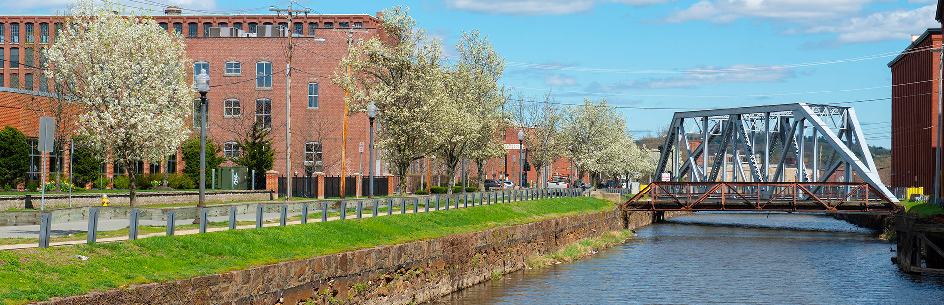 Pemberton Mill Bridge on the Merrimack River North Canal at Lawrence Heritage State Park in downtown Lawrence, Massachusetts MA, USA. The bridge is a steel truss bridge with a pedestrian walkway and a view of the river and city skyline. The image is taken during the day with clear skies and vibrant colors, showcasing the historic architecture and natural beauty of the area.