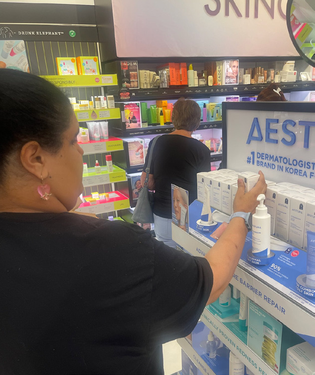 A woman with dark hair pulled back is wearing a black long-sleeved top. She is in a retail environment, reaching toward a product display shelf that contains skincare items while a coworker browses.